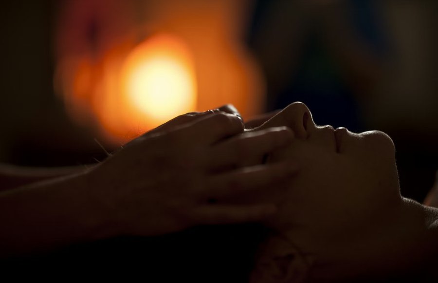 Reiki session — gentle hands on forehead and temple in a calm, dimly lit setting; person at rest during therapeutic touch
